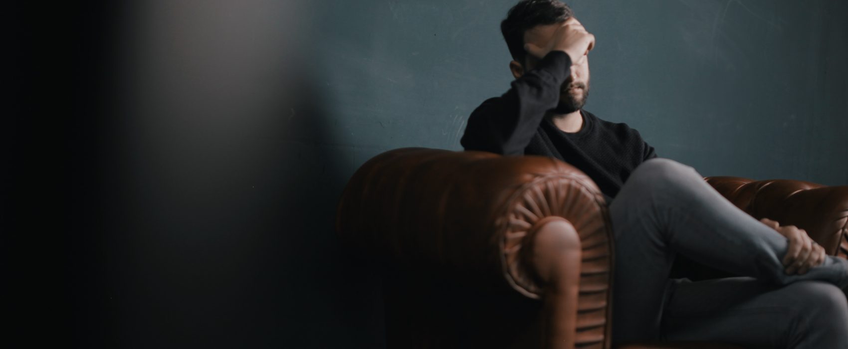 A male employee who is experiencing workplace bullying sits alone on a sofa with his hands over his eyes.